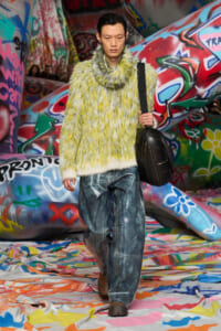 Model walks the runway in a neon yellow-green fuzzy sweater, carrying a black round shoulder bag against a graffiti-covered backdrop.