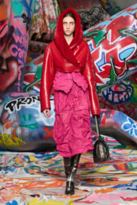 Model wearing a red oversized hooded jacket over a bright pink crinkled skirt, walking on a graffiti-covered runway with a black handbag.