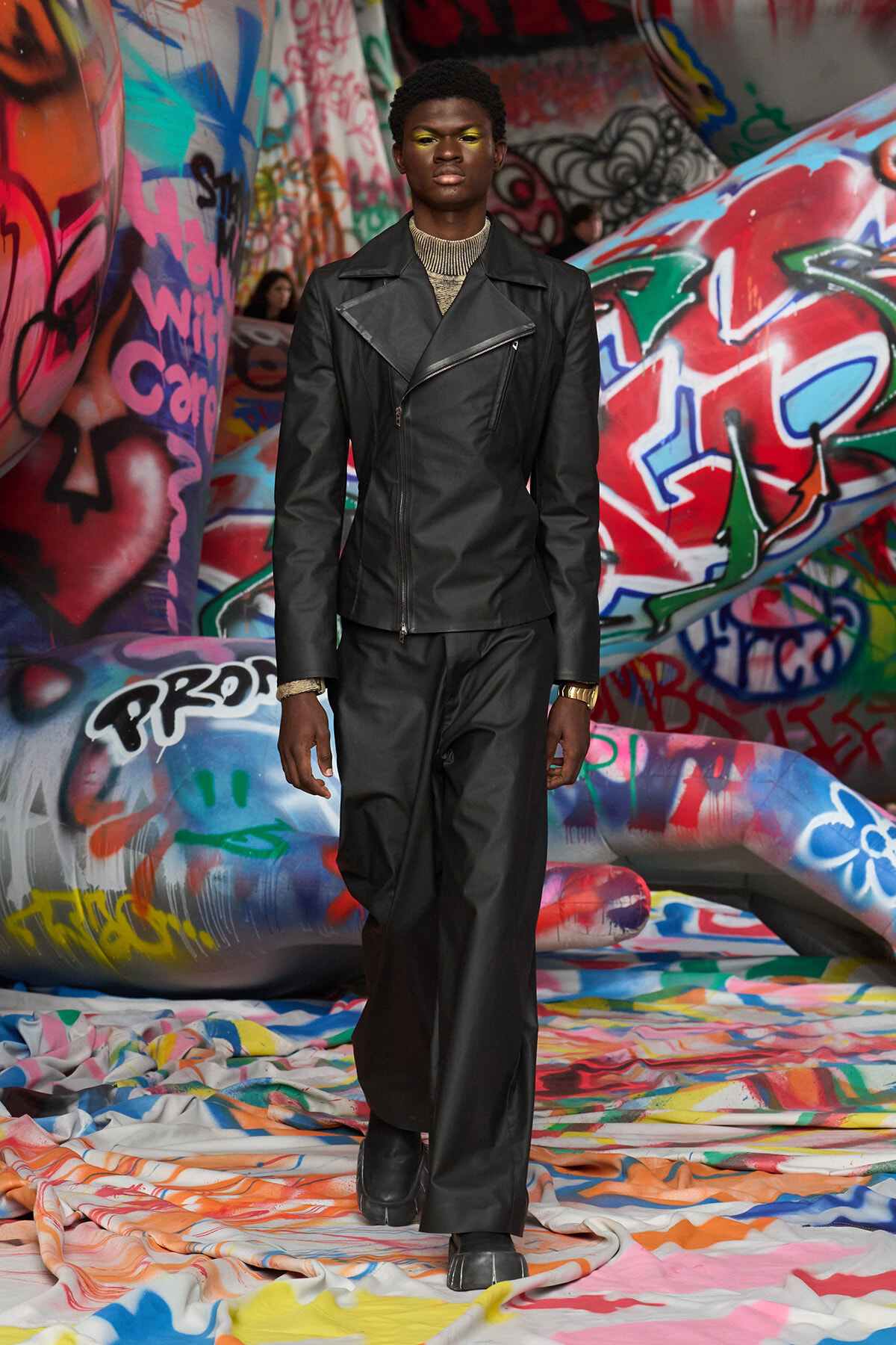 Model wearing a black leather jacket and matching pants walks forward in a graffiti-covered tunnel.