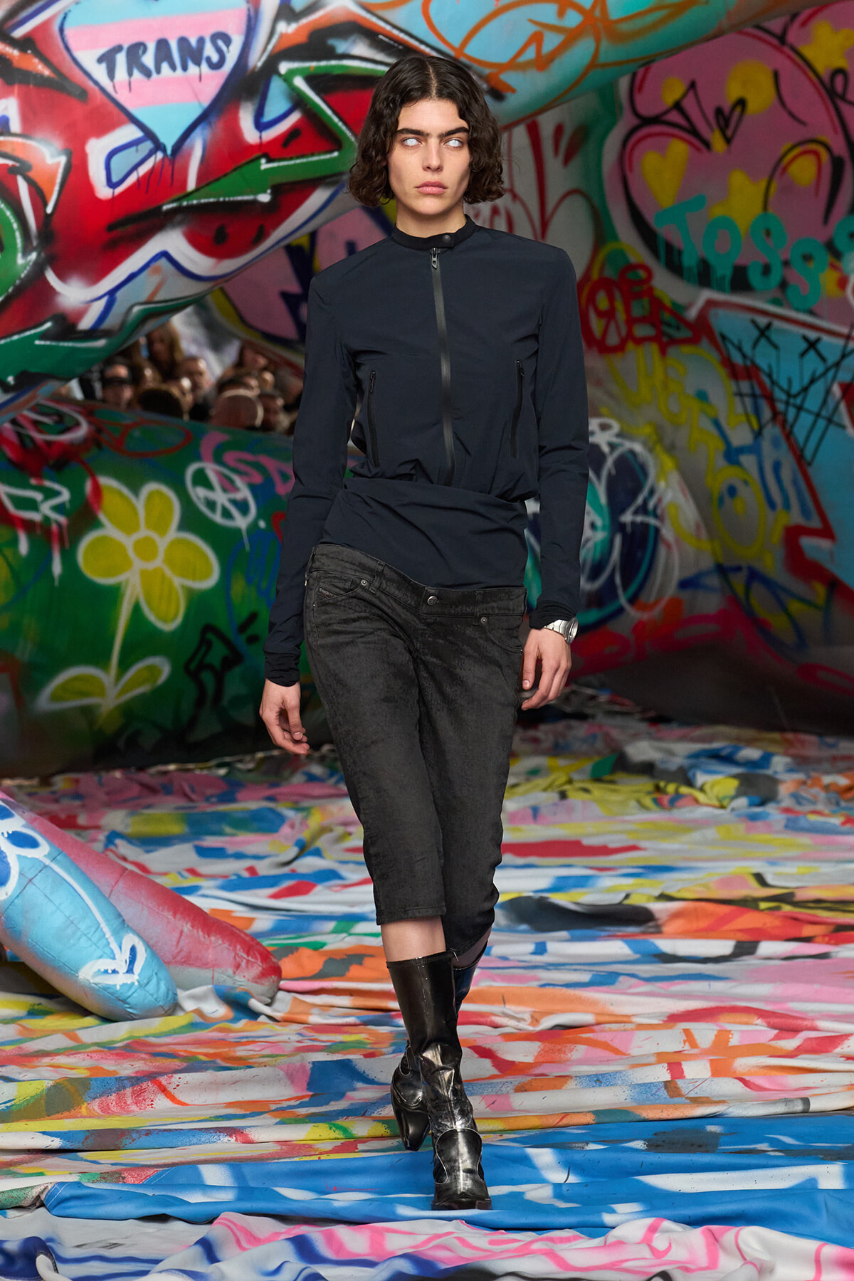 Model walks a runway in a navy zip-front jacket, black cropped jeans, and black boots, with a graffiti backdrop.