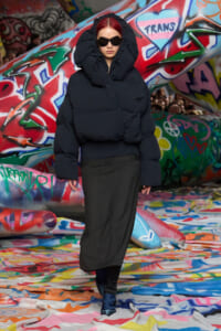 Person in an oversized black puffer jacket with hood and dark sunglasses stands in a graffiti-covered tunnel.