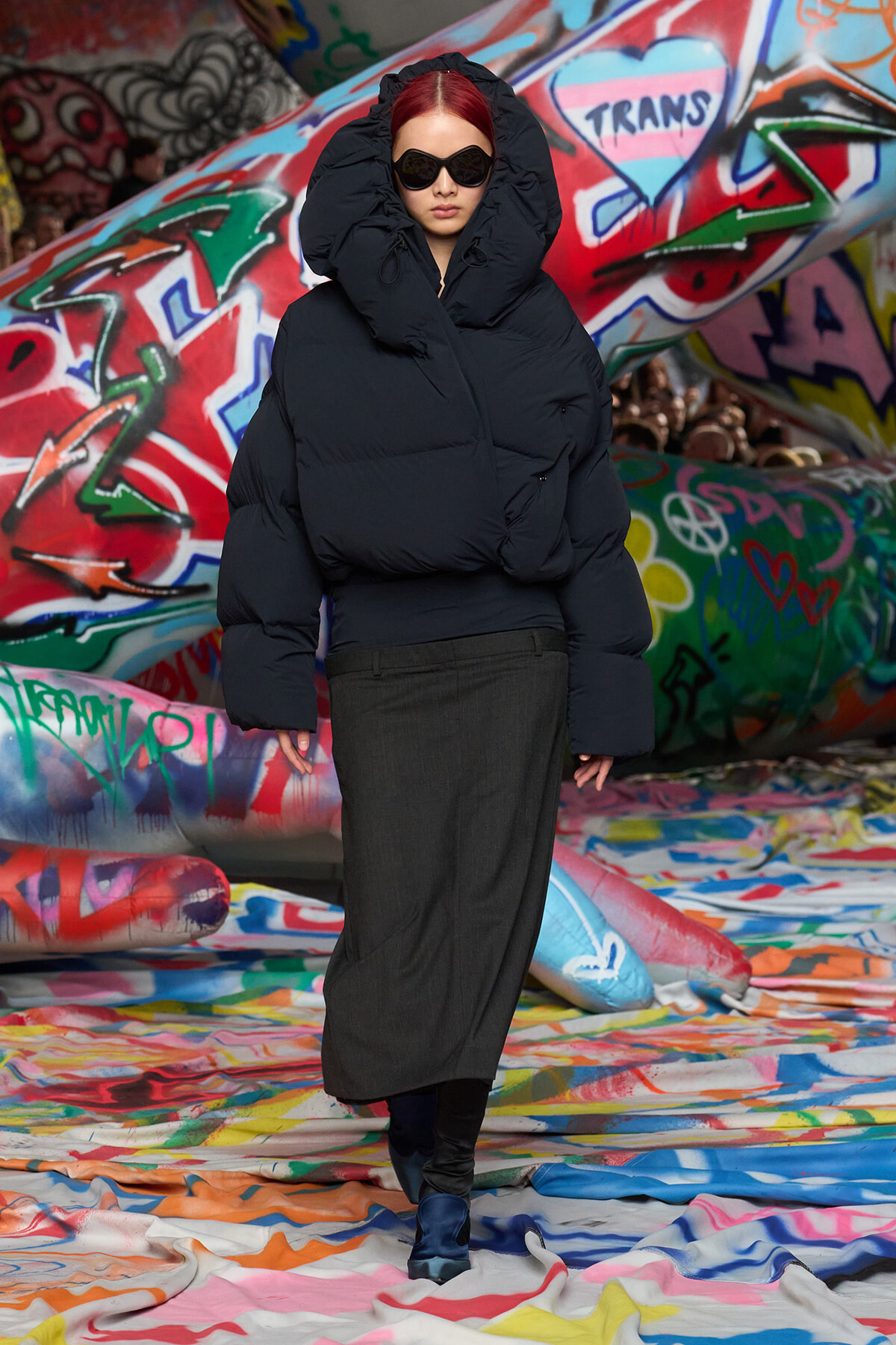 Person in an oversized black puffer jacket with hood and dark sunglasses stands in a graffiti-covered tunnel.