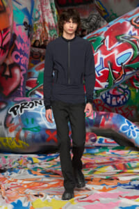 Male model wearing a dark zip jacket and black pants walks along a graffiti-covered runway set in a fashion show.](https://)
