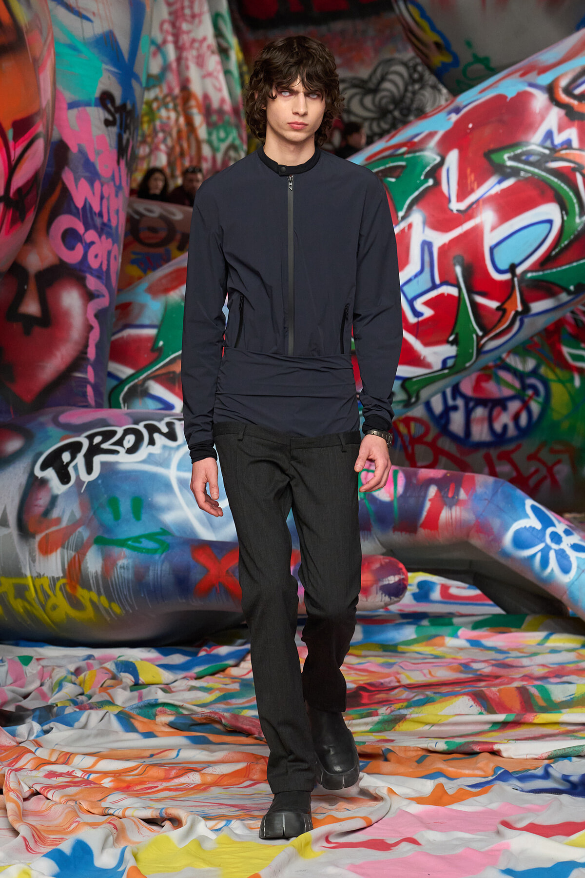 Male model wearing a dark zip jacket and black pants walks along a graffiti-covered runway set in a fashion show.](https://)