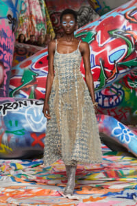 Model wearing a fringed beige textured dress posing in front of colorful graffiti walls.