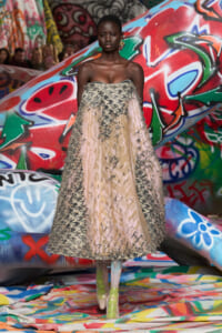 Model in a strapless beige gown with textured appliqué walking on a graffiti-covered runway backdrop.