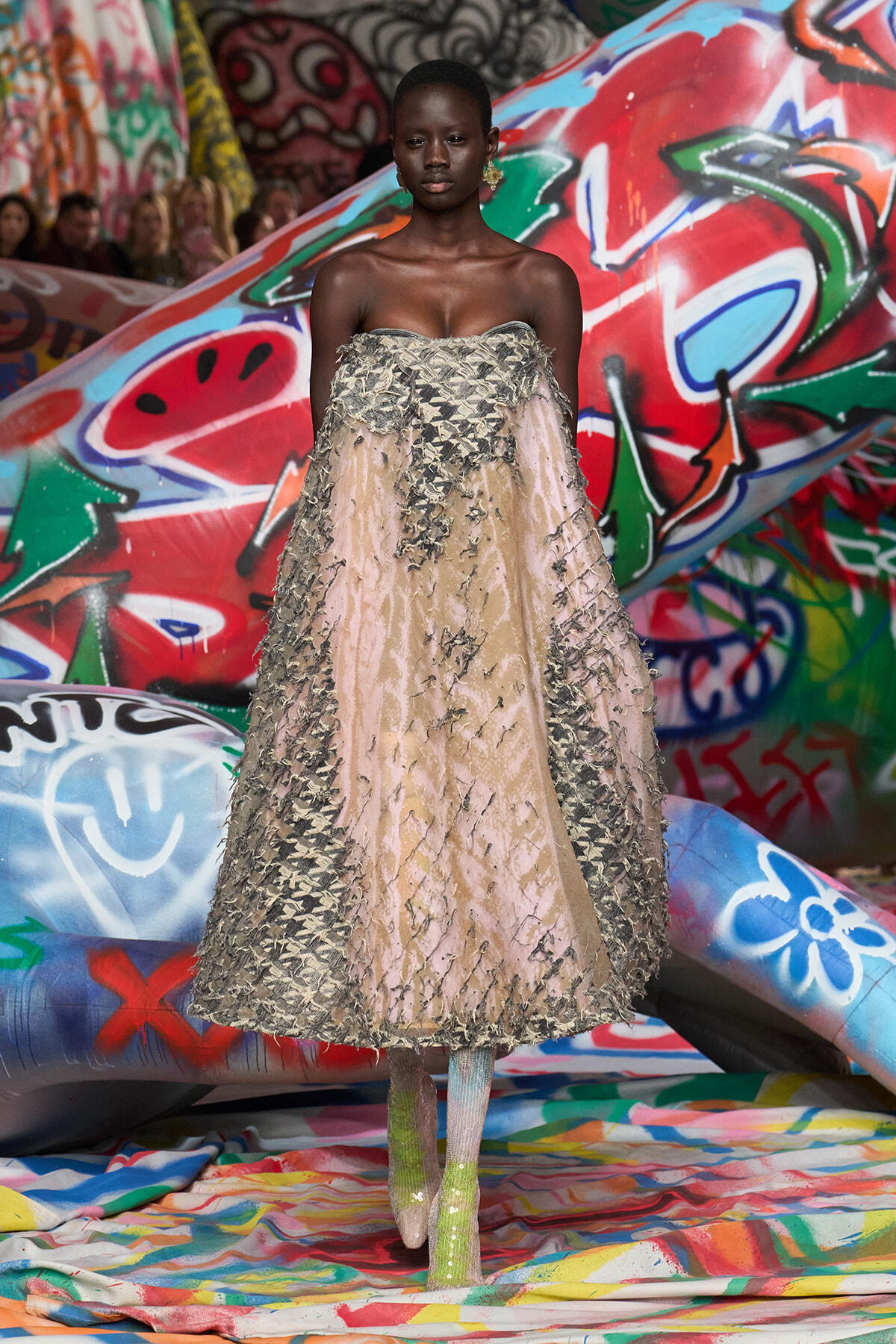 Model in a strapless beige gown with textured appliqué walking on a graffiti-covered runway backdrop.