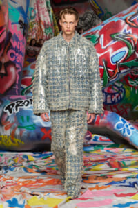 Model wears a silver, fringed textured suit walking a graffiti-covered runway scene.