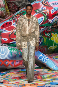Male model in a metallic beige textured suit walking a graffiti-covered runway during a fashion show.