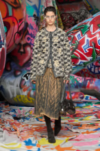 Model wearing a textured beige-black cardigan over a tiger-print skirt stands in a graffiti-covered tunnel with colorful fabric floor.