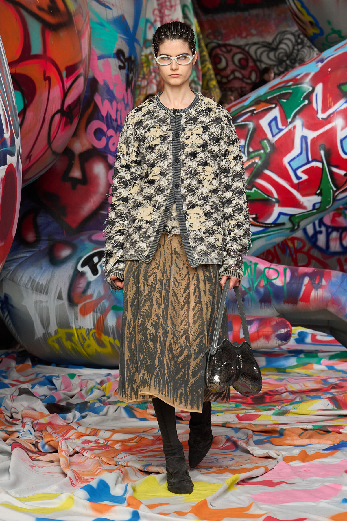 Model wearing a textured beige-black cardigan over a tiger-print skirt stands in a graffiti-covered tunnel with colorful fabric floor.