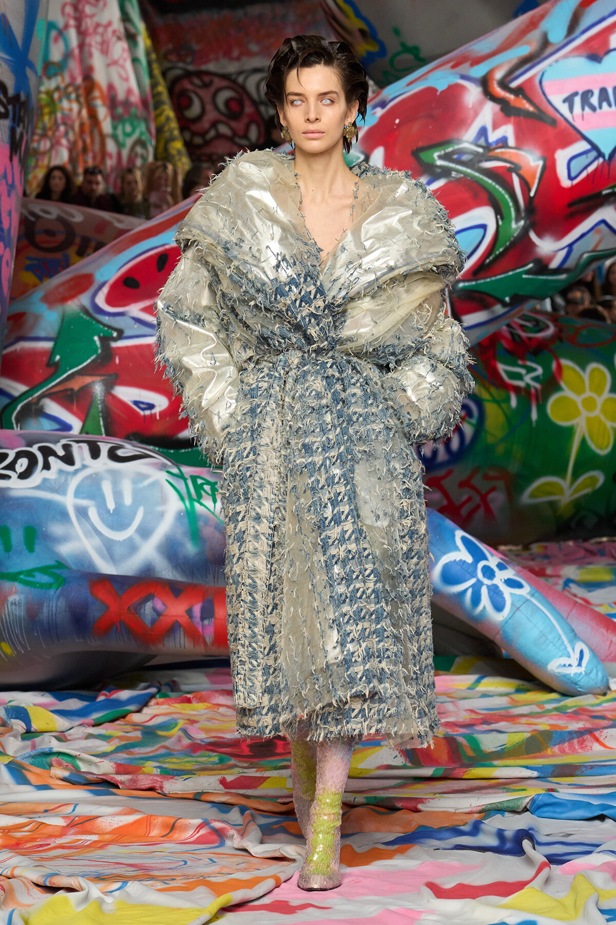 Model in an oversized metallic silver coat with fringe detailing, posing on a graffiti-covered runway backdrop.