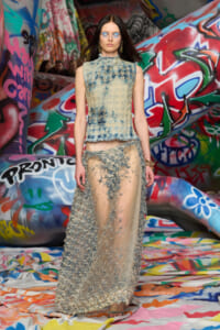 Fashion model on a graffiti-covered runway wearing a textured beige-blue sleeveless top and a sheer, embroidered skirt.