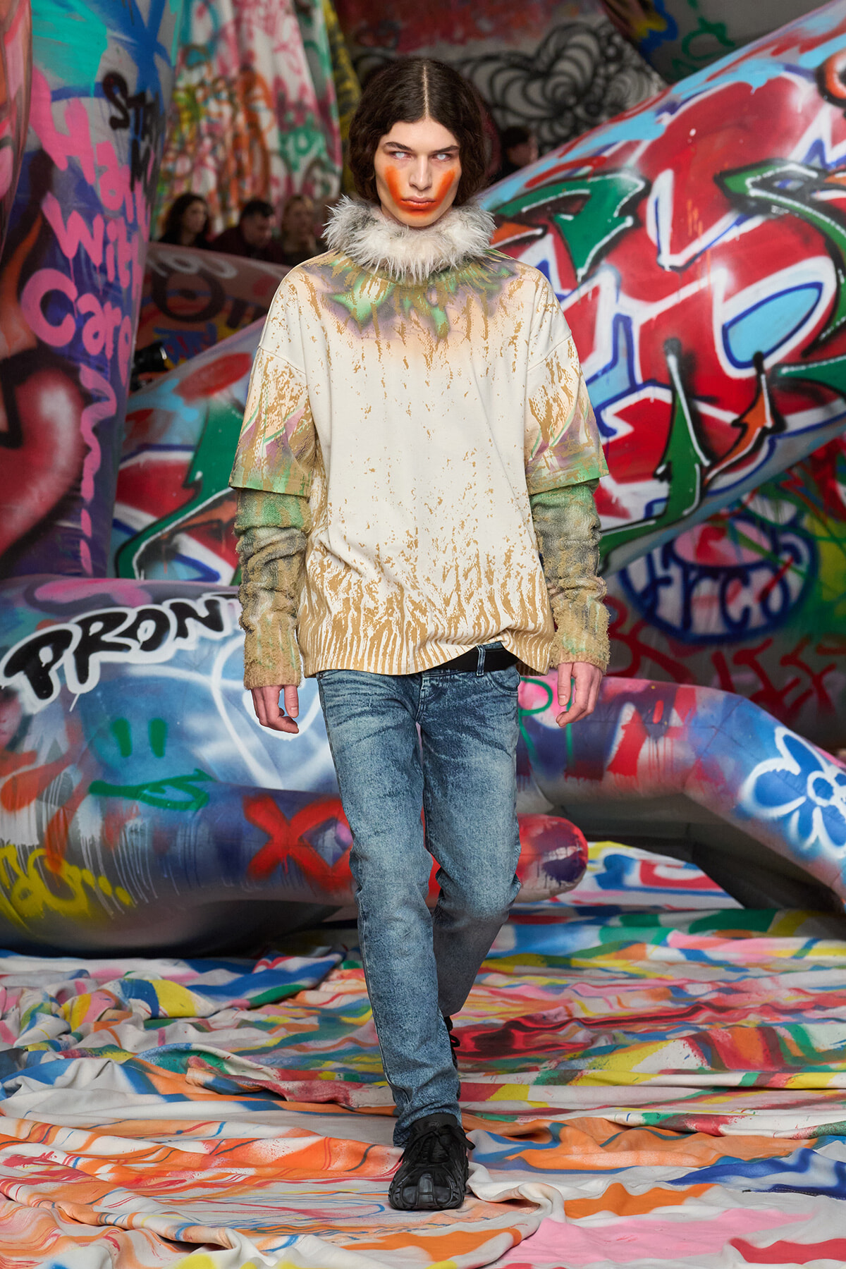 Model walking a colorful graffiti-lined runway in a beige paint-splattered top with feathered collar and blue jeans, black shoes.