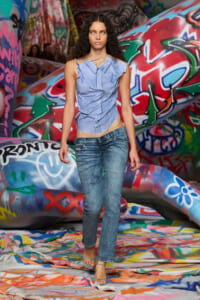 Model walking on a graffiti-covered runway wearing a light blue draped sleeveless top, mid-rise faded jeans, and strappy sandals.