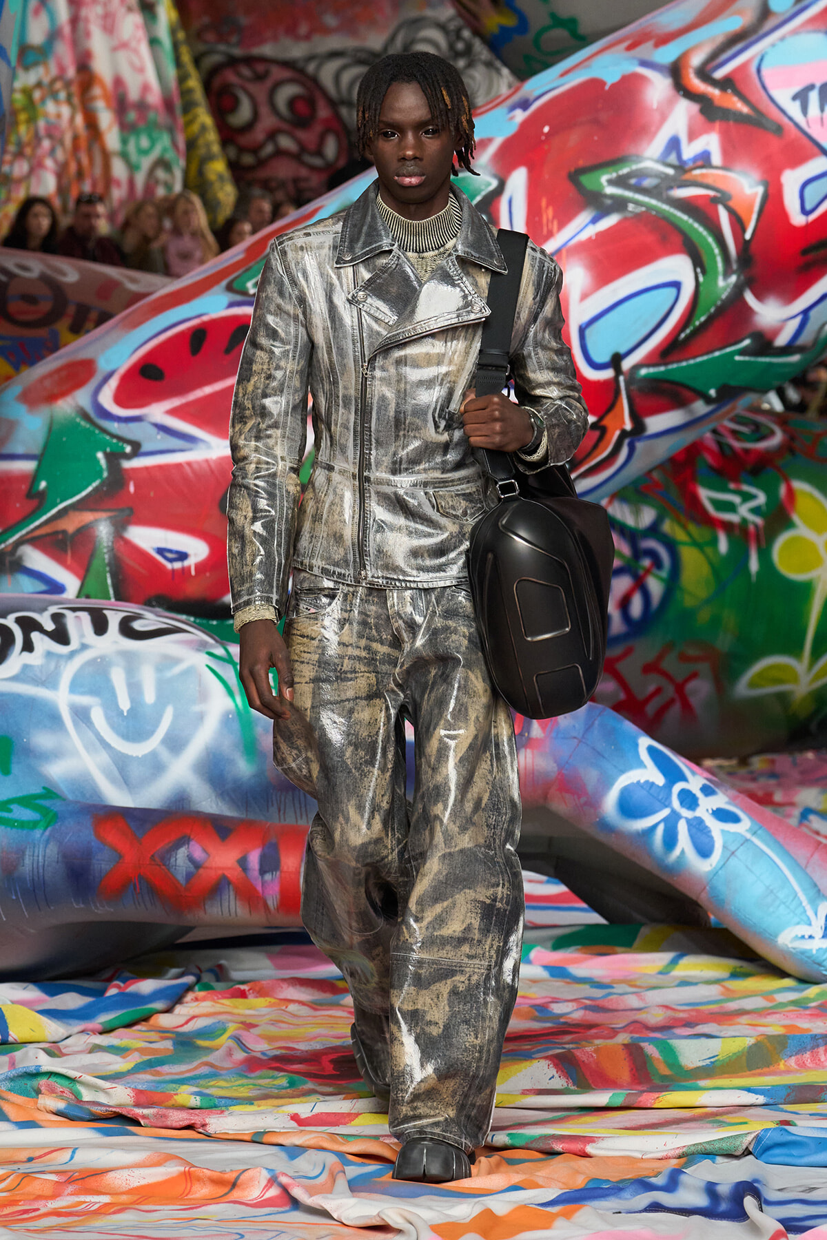 Male model in a shiny silver metallic suit and black crossbody bag walks past colorful graffiti-filled backdrop.