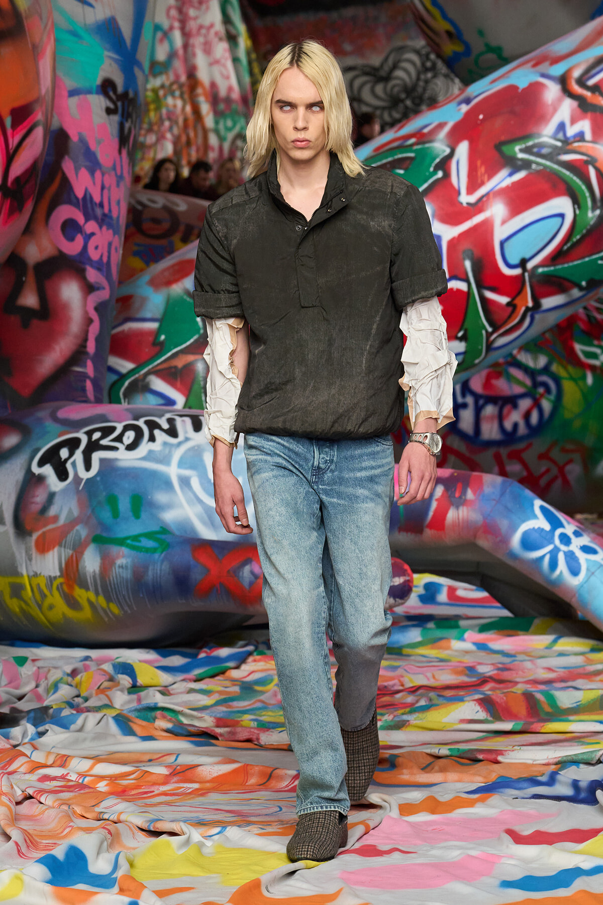 Male fashion model with shoulder-length blond hair walks through a graffiti-covered tunnel, wearing a dark shirt over scrunched white sleeves and light jeans.