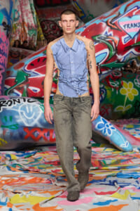 Male model walks a graffiti-covered runway wearing a light blue ripped shirt and distressed gray jeans.