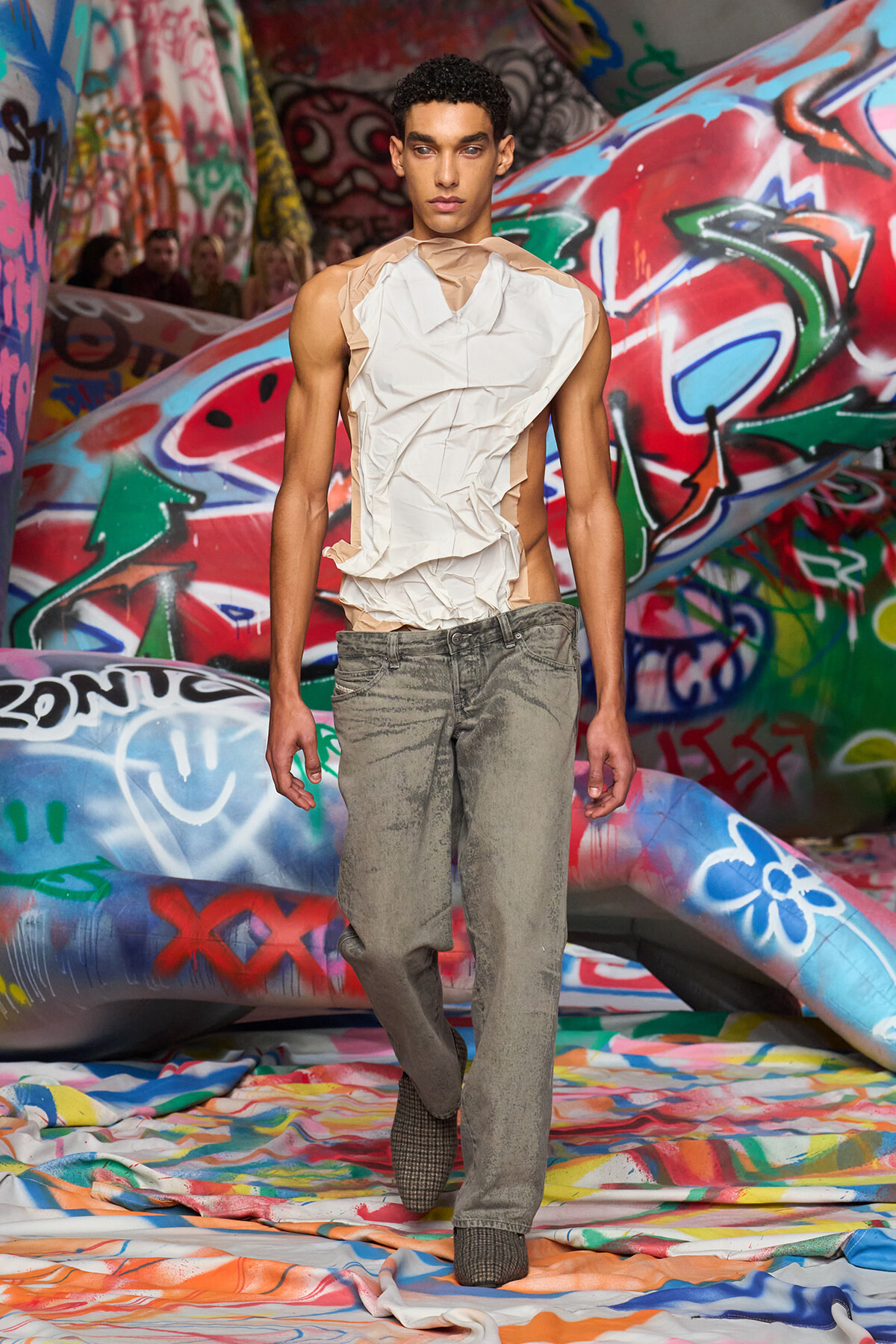 Male model on a runway wearing a crumpled beige sleeveless top and gray denim jeans, with a colorful graffiti backdrop.