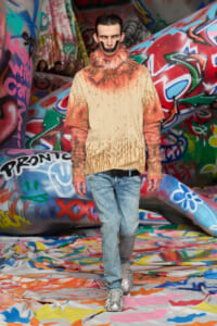 Model wearing a beige spray-paint-themed knit top with orange fur collar and orange sleeves, paired with light-wash jeans on a graffiti-covered runway.