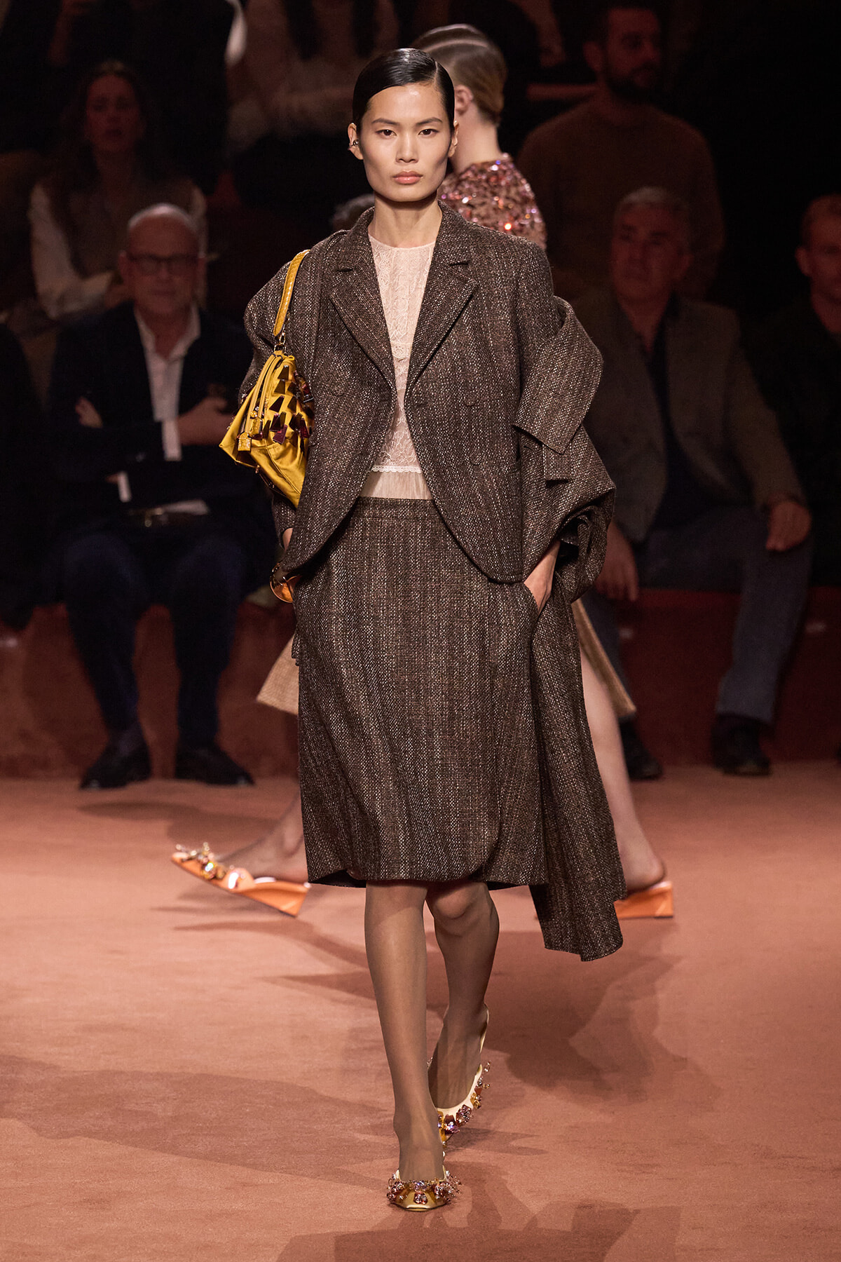 Model walking a runway in a brown textured suit with an oversized coat and a yellow handbag slung over the shoulder, beige top underneath.