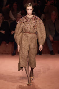 Model on a runway wearing a beige textured coat with crystal embellishments on the chest and shoulders, belted at the waist.