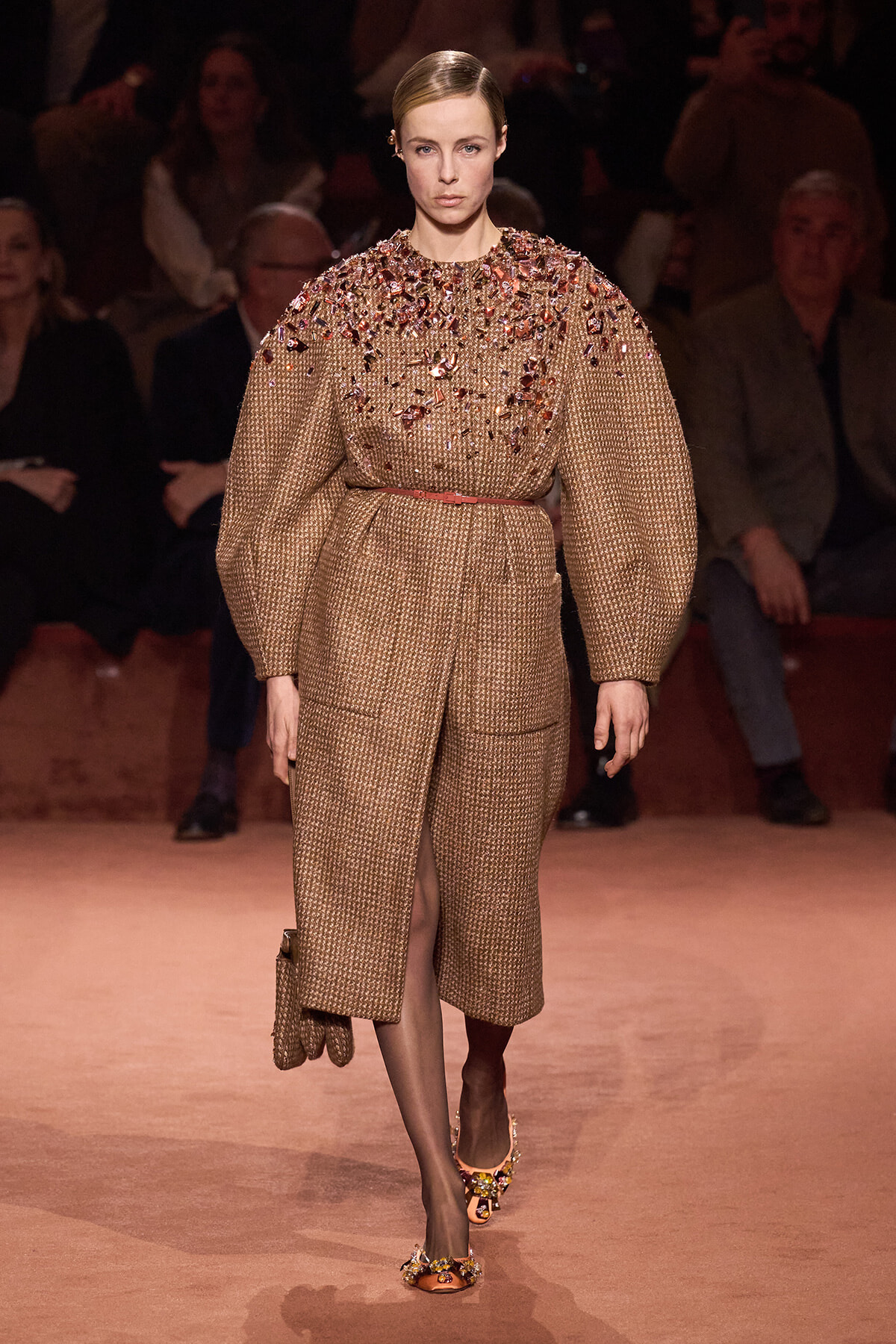 Model on a runway wearing a beige textured coat with crystal embellishments on the chest and shoulders, belted at the waist.