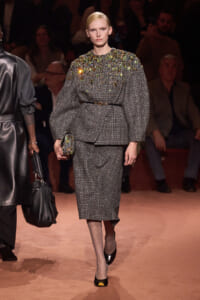 Model walks the runway in a gray plaid belted blazer with oversized sleeves and a matching midi skirt, embellished with jeweled details on the shoulders, while holding a small jeweled clutch.
