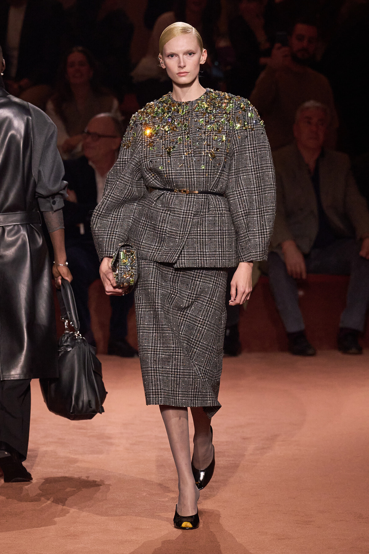 Model walks the runway in a gray plaid belted blazer with oversized sleeves and a matching midi skirt, embellished with jeweled details on the shoulders, while holding a small jeweled clutch.