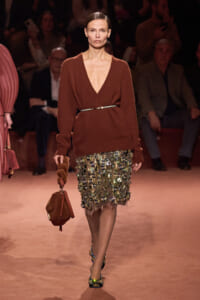 Runway model in a rust brown V-neck knit sweater belted at the waist, with a jeweled metallic skirt and a brown handbag in hand.