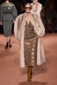 Model on a runway wearing a long cream fur coat, a brown jeweled-knit top, and a metallic gold patterned skirt with tan boots and amber sunglasses.