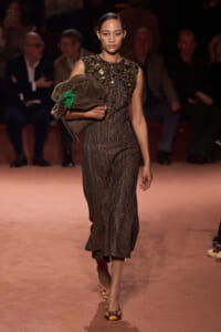 Model walks the runway in a sparkly sleeveless top and shimmering wide-leg pants, carrying a brown coat with a green accessory.