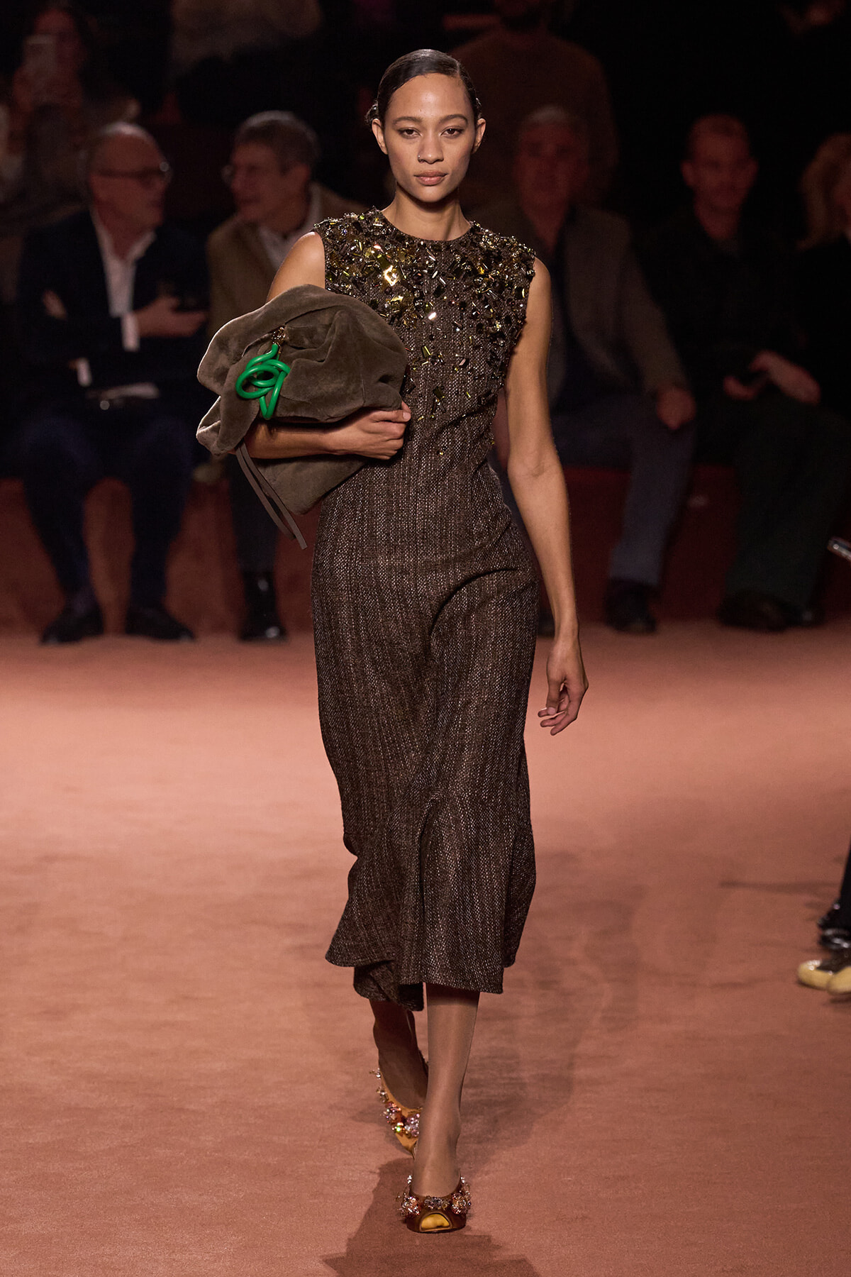 Model walks the runway in a sparkly sleeveless top and shimmering wide-leg pants, carrying a brown coat with a green accessory.