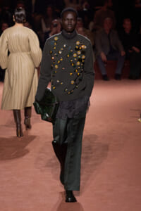 Model on a runway wearing a dark green embellished sweater with jewel appliqués, lace hem, dark green satin trousers, and carrying a glossy emerald clutch.