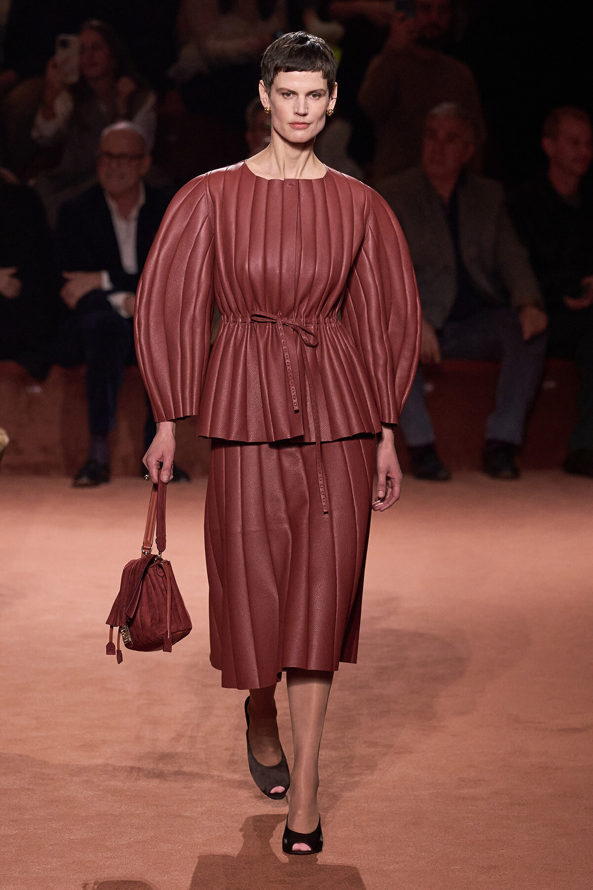 Female model walking the runway in a mauve pleated leather ensemble with a matching belt and handbag.