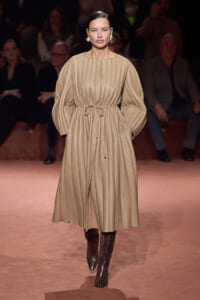 Model walking the runway in a long beige pleated dress with gathered waist and voluminous sleeves, paired with leopard print boots.