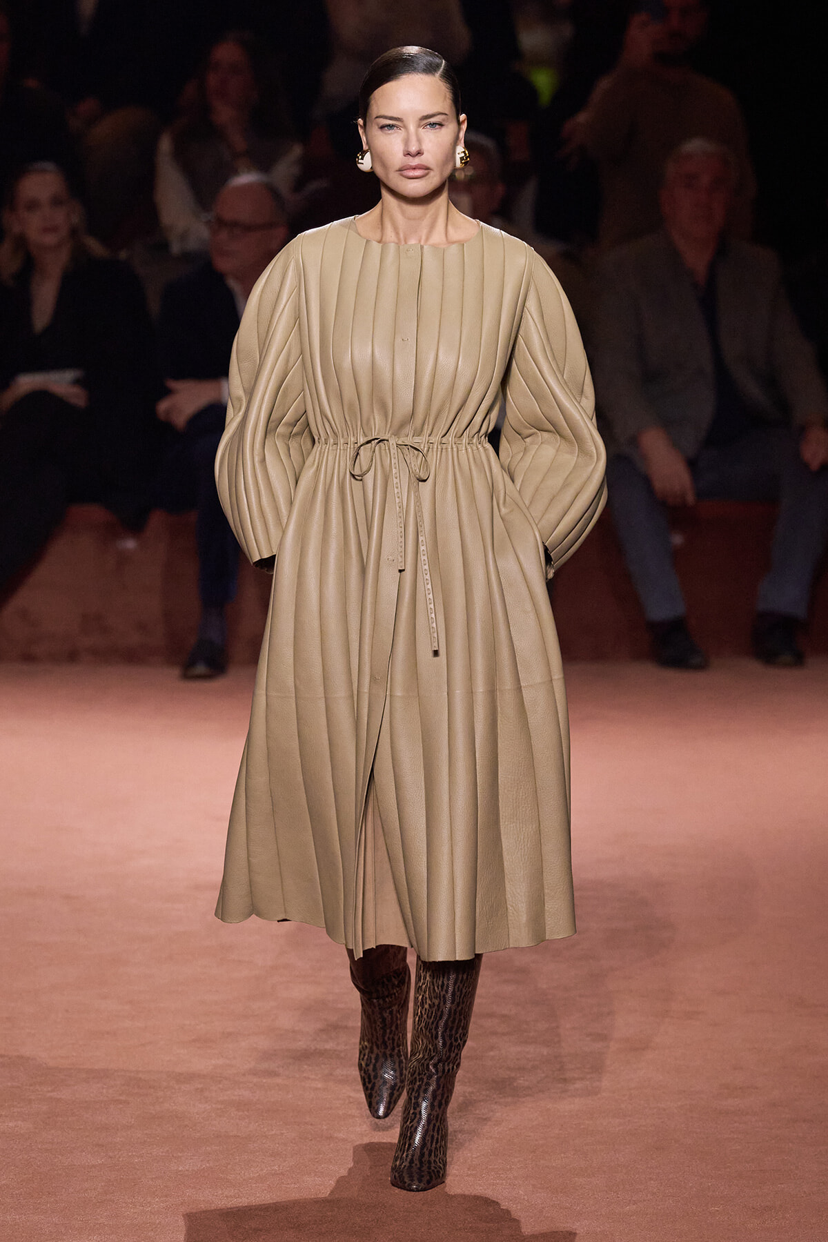 Model walking the runway in a long beige pleated dress with gathered waist and voluminous sleeves, paired with leopard print boots.