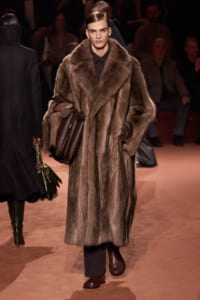 Model walks the runway in a full-length brown fur coat, black outfit, and a brown leather bag.