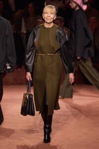 Model walks the runway in an olive dress with black leather sleeves, a slim belt, knee-high boots, and a handbag.