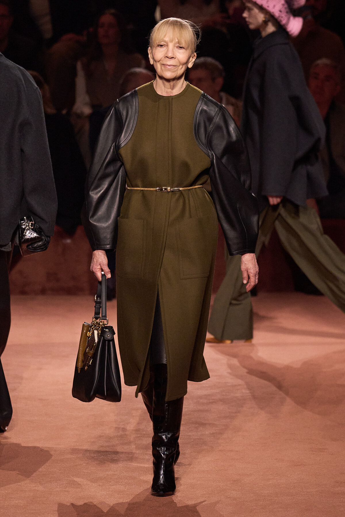 Model walks the runway in an olive dress with black leather sleeves, a slim belt, knee-high boots, and a handbag.