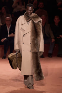 Model walks the runway wearing a beige double-breasted wool coat with a fur scarf draped over the right shoulder, carrying a matching fur-trimmed bag.