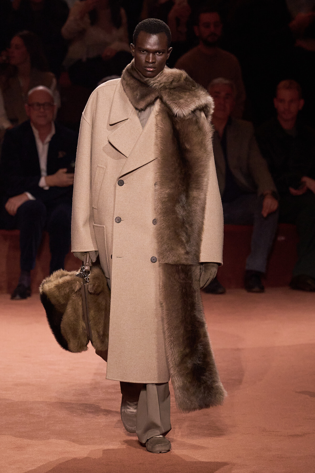 Model walks the runway wearing a beige double-breasted wool coat with a fur scarf draped over the right shoulder, carrying a matching fur-trimmed bag.