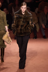 Model on a runway wearing a dark brown fur coat and black wide-leg pants, serious expression, audience in the background.