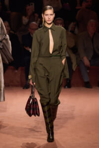Model walks a fashion runway in an olive-green jumpsuit with a front cutout, belt, and knee-high striped boots, carrying a dark burgundy handbag. Audience is in the background.