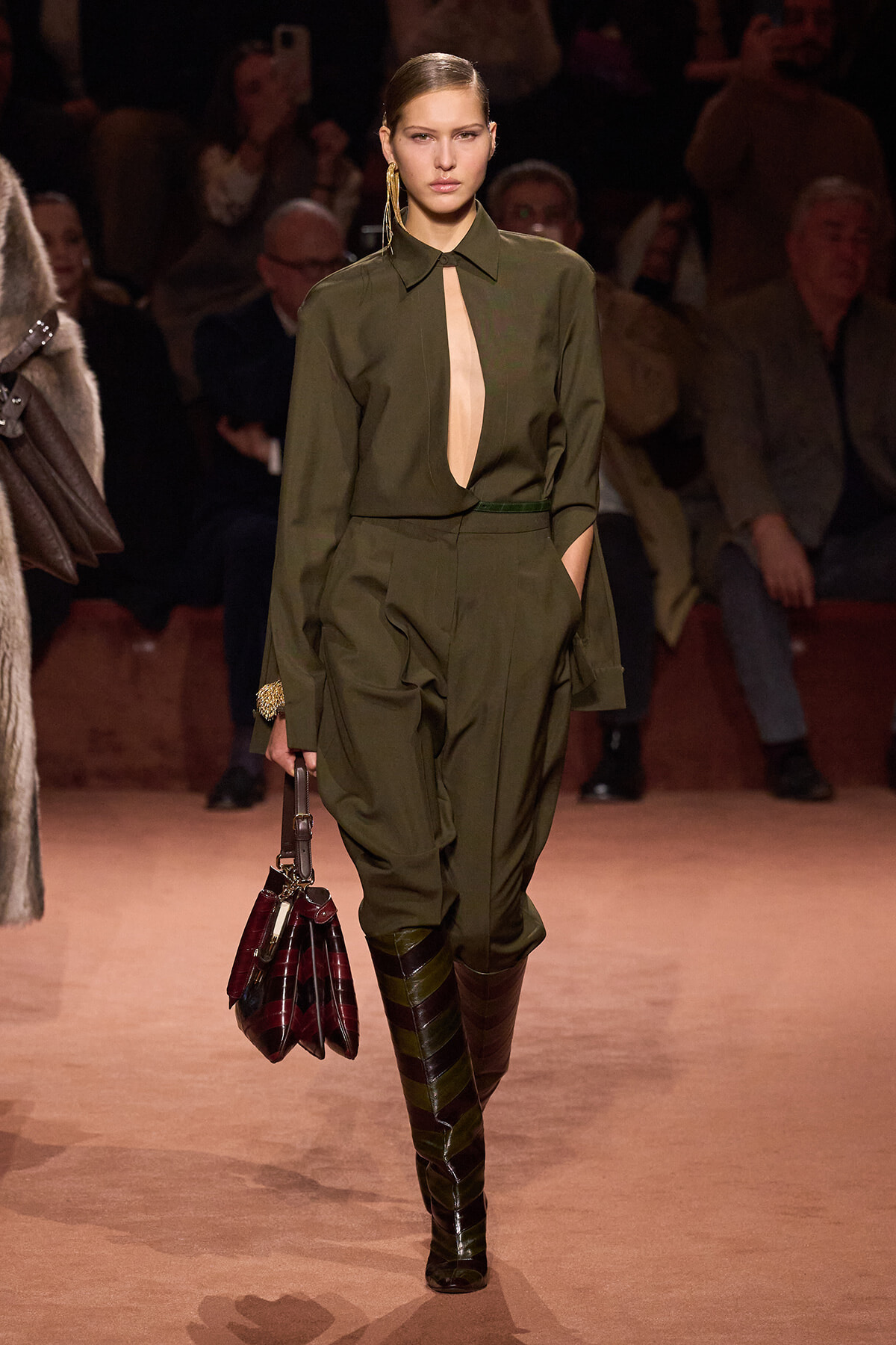 Model walks a fashion runway in an olive-green jumpsuit with a front cutout, belt, and knee-high striped boots, carrying a dark burgundy handbag. Audience is in the background.