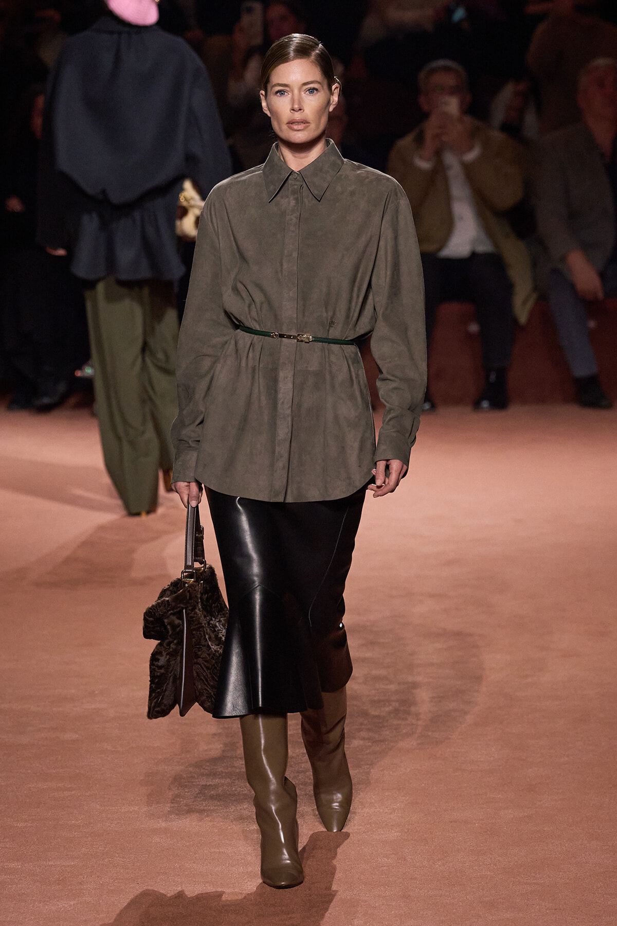 Model walking a runway in an olive shirt-dress with a thin belt, paired with a black midi leather skirt and tan boots, carrying a fur handbag.