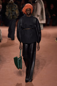 Model walks the runway in a black leather-trim ensemble with a bright orange knitted hat and a green handbag.