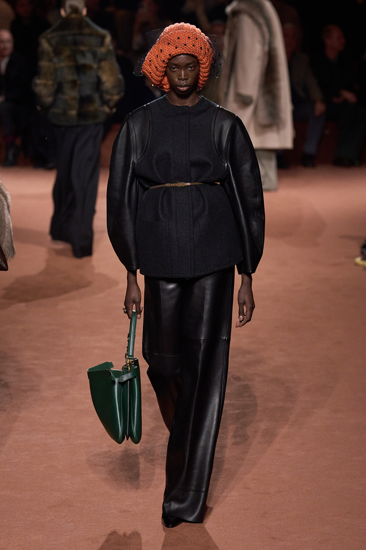 Model walks the runway in a black leather-trim ensemble with a bright orange knitted hat and a green handbag.