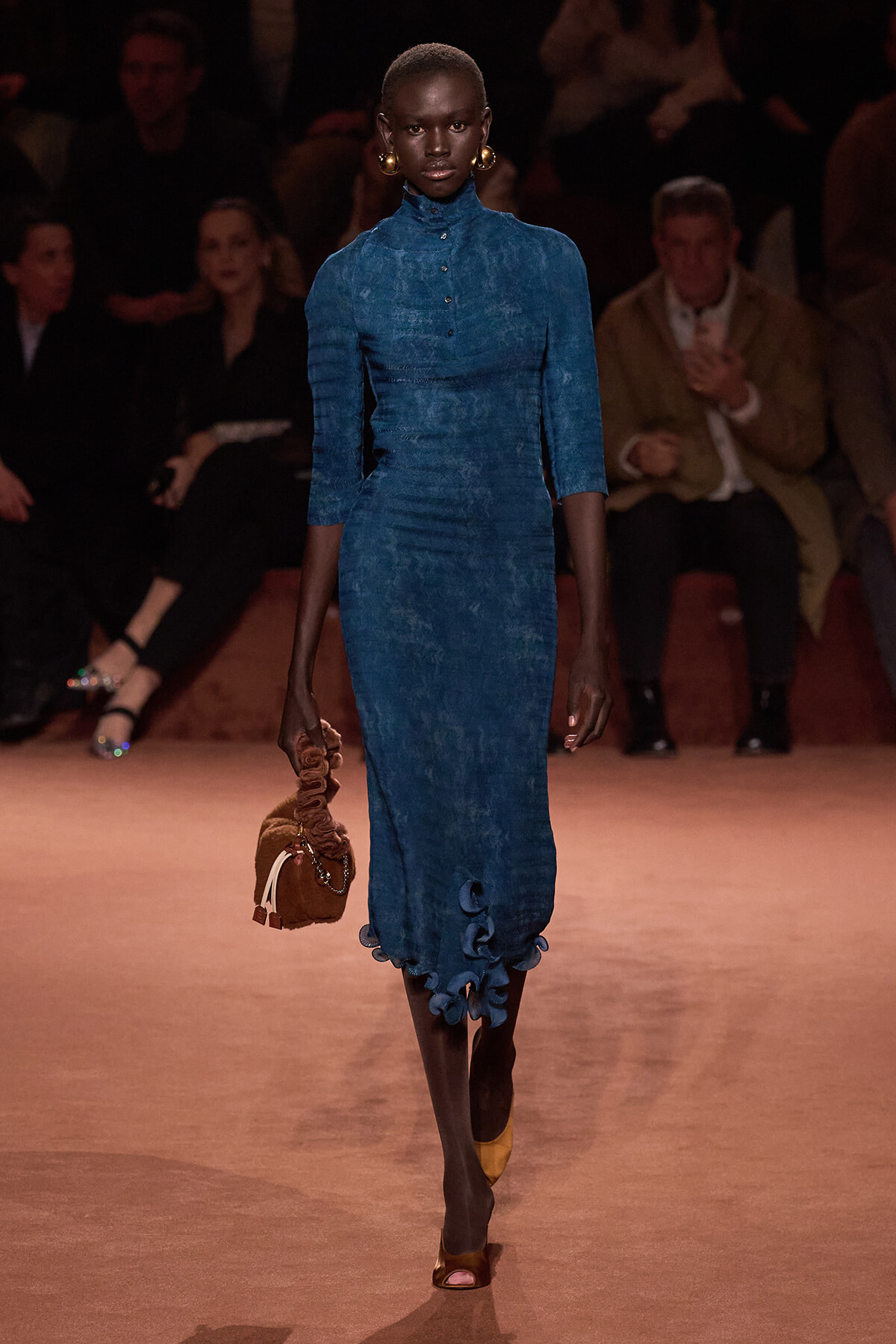 Model walking the runway in a blue, knee-length textured dress with three-quarter sleeves and ruffled hem; gold hoop earrings, yellow open-toe heels, and a brown gathered handbag.