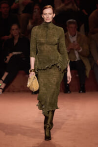 Model walks the runway in an olive green, textured dress with a high neckline and ruffled peplum, carrying a tan handbag; audience in dim light behind.